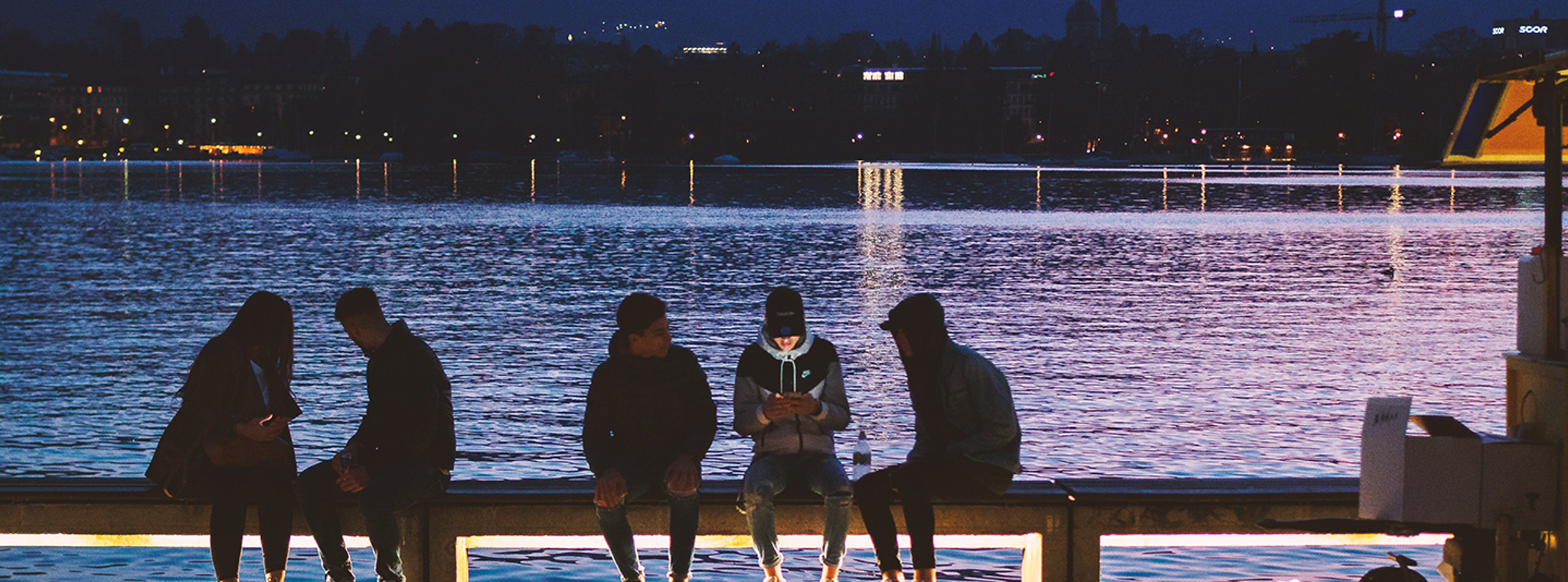 A photo of group of people sat by a lake using their mobile phones