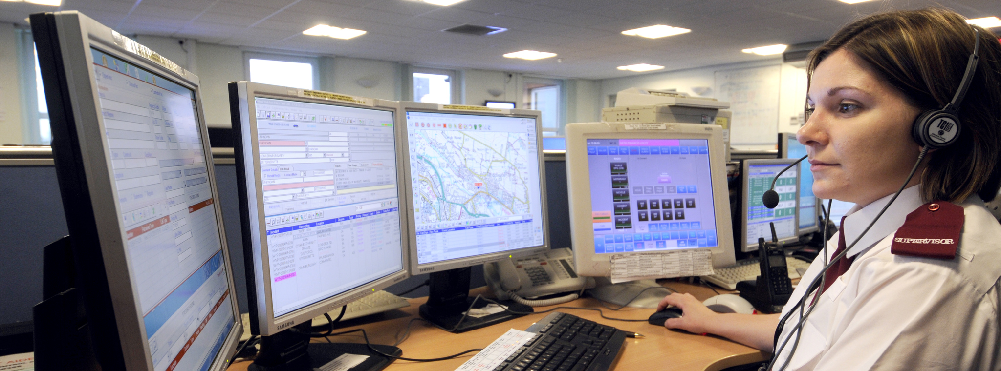 A lady in a police control room answering a call with four PC monitors around her