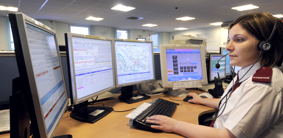 A lady in a police control room answering a call with four PC monitors around her