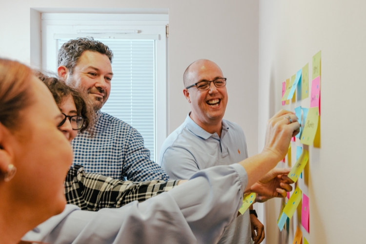 A group of people smiling and adding Post-It notes to a whiteboard