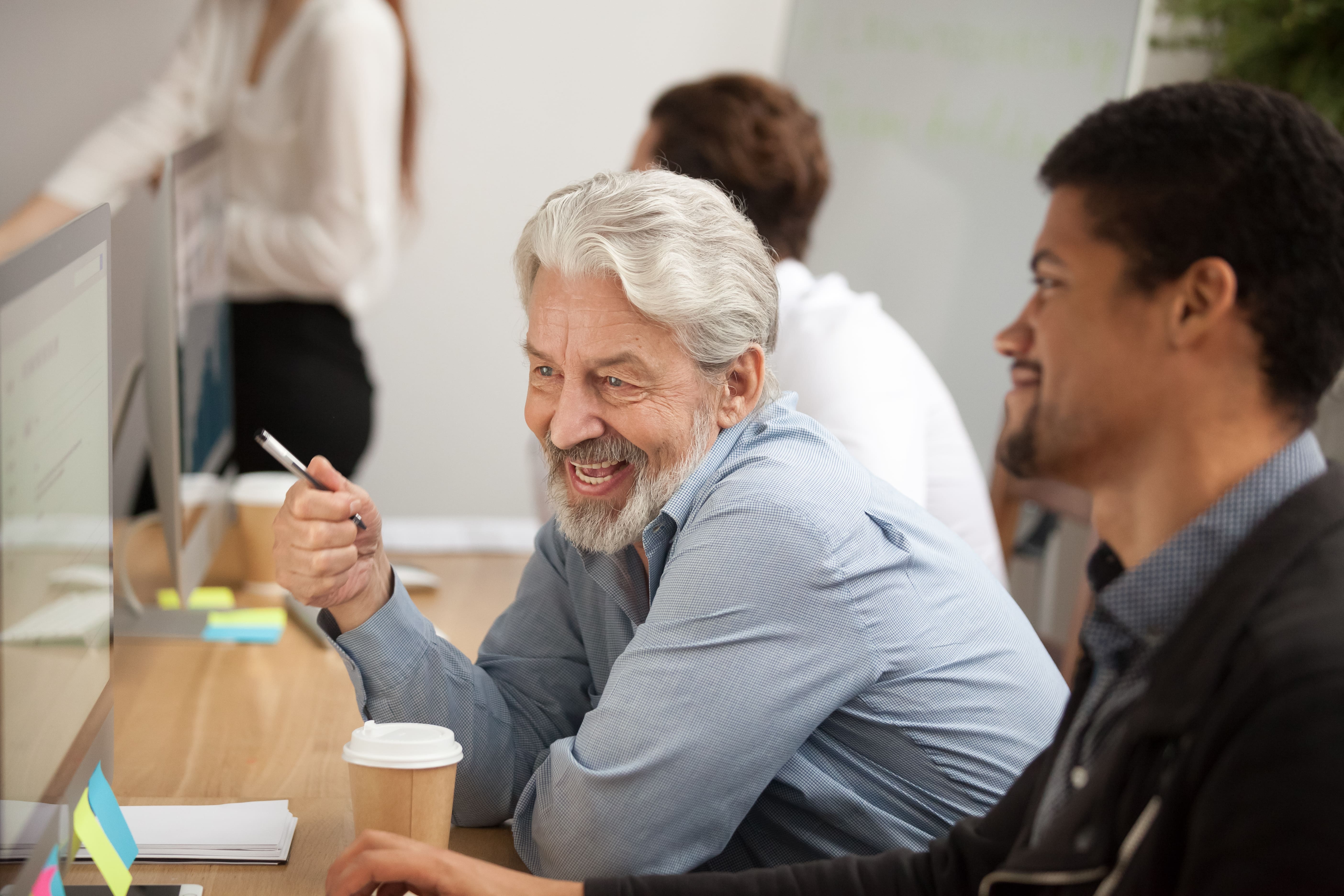 Senior man with a coffee cup sat next to a young man working on computer